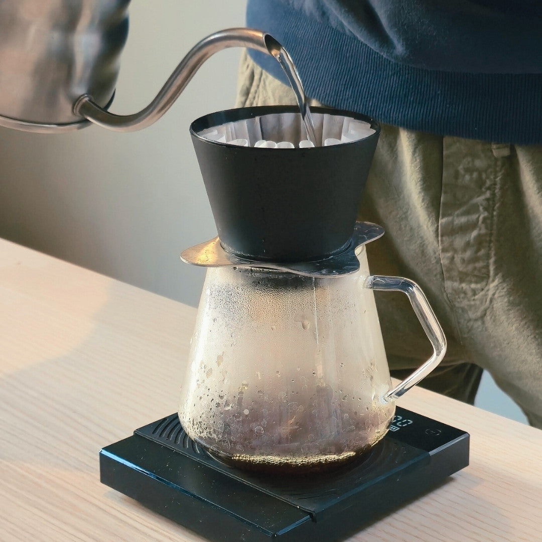 Person pouring water into a coffee filter on a clear glass carafe with a black base.