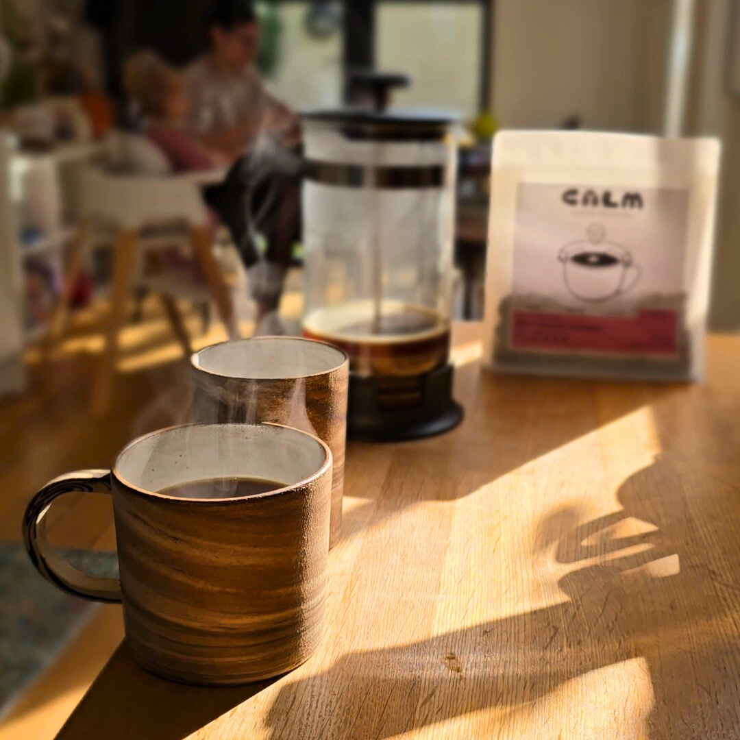 Two steaming mugs of Calm Coffee with French press and specialty coffee bag in soft morning light, family in background