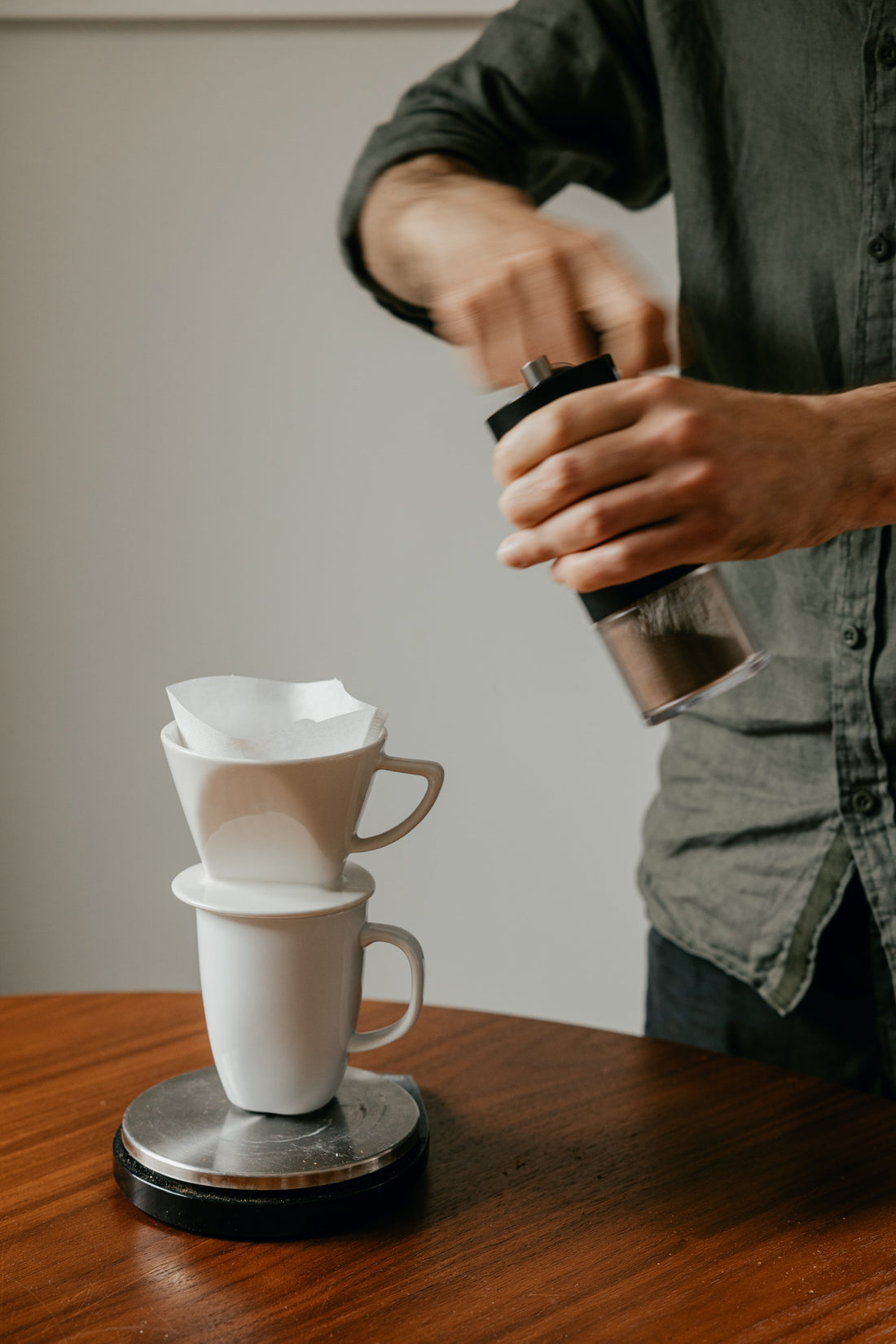 A man hand grinding coffee next to a coffee filter cone sitting on mug and scales.