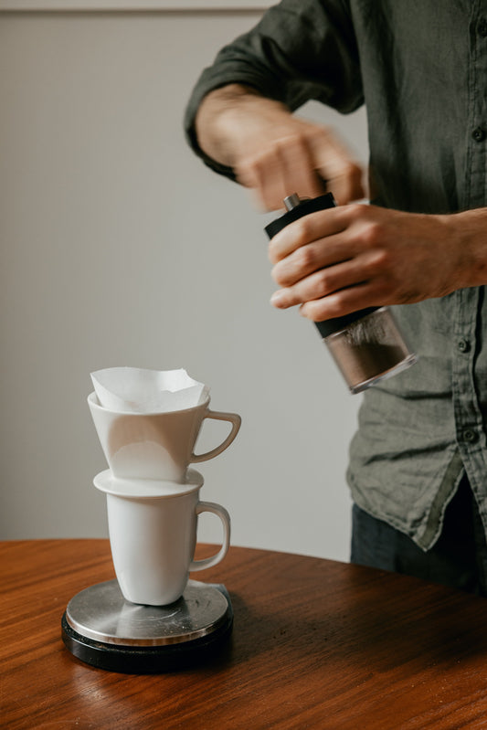 A man hand grinding coffee next to a coffee filter cone sitting on mug and scales.