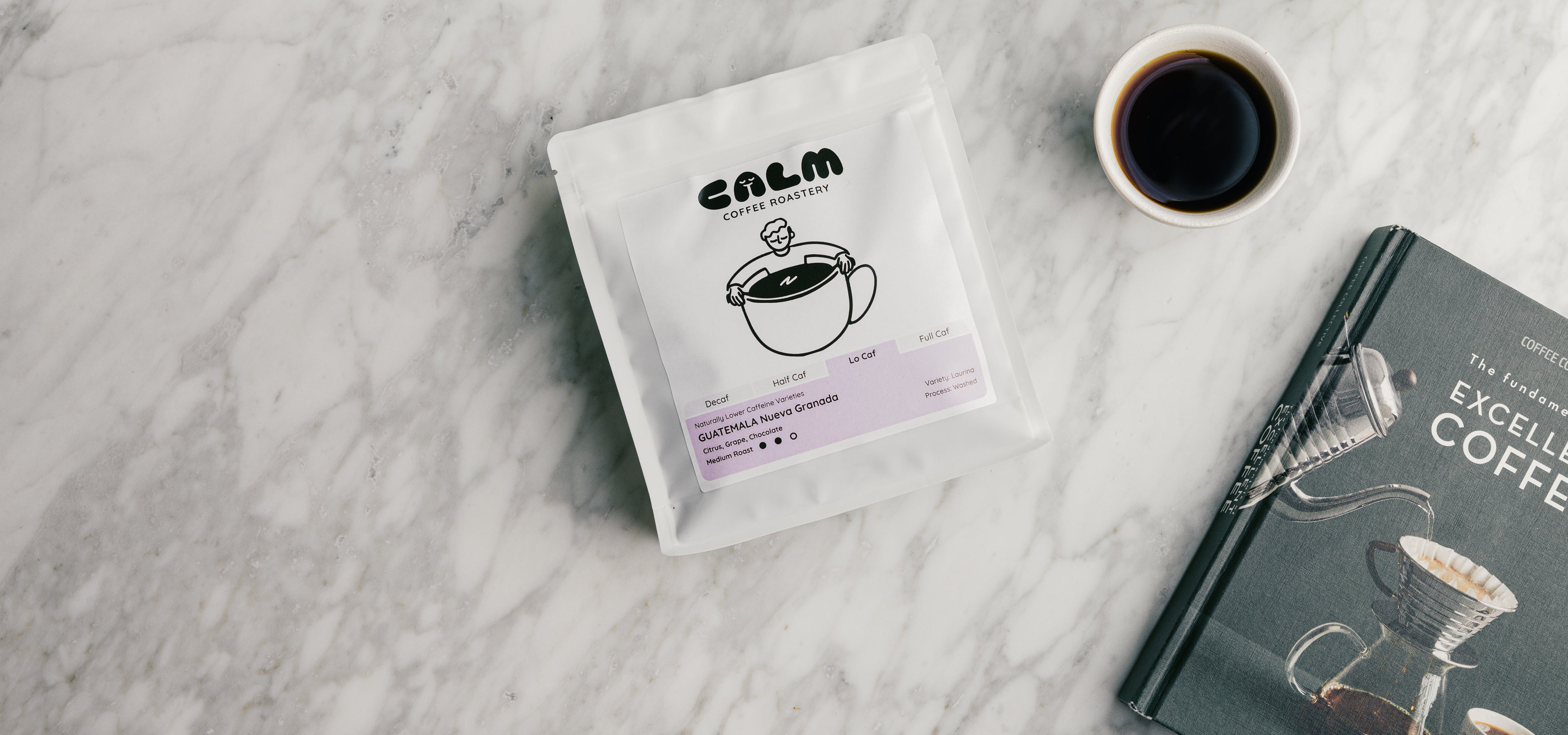 Calm Coffee Roastery Low Caf Guatemala Nueva Granada bag on marble with coffee cup and coffee book