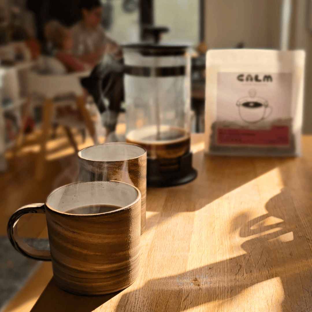 Two mugs with steaming coffee on a wooden table, with a French press and coffee bag in the background.