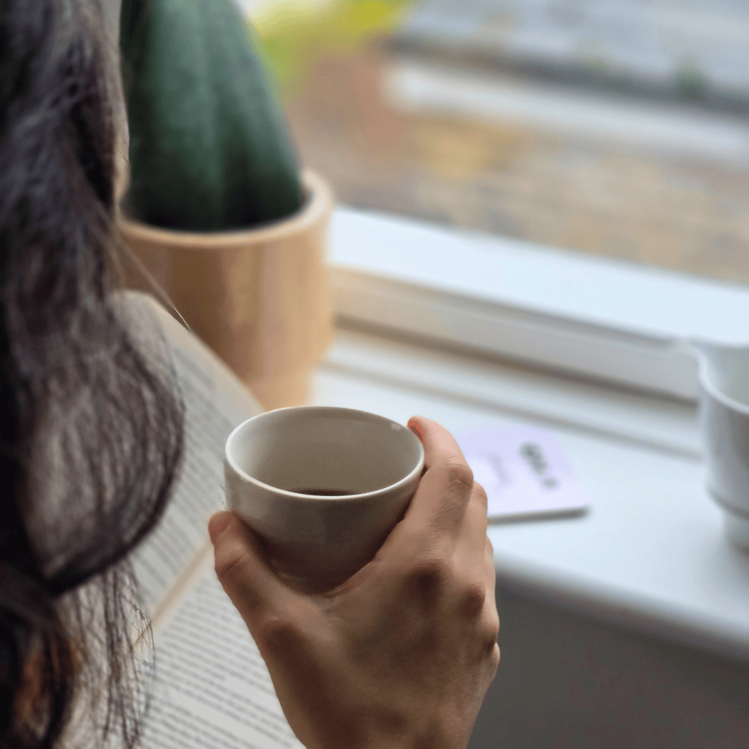 Person holding a white coffee mug by a window while reading a book